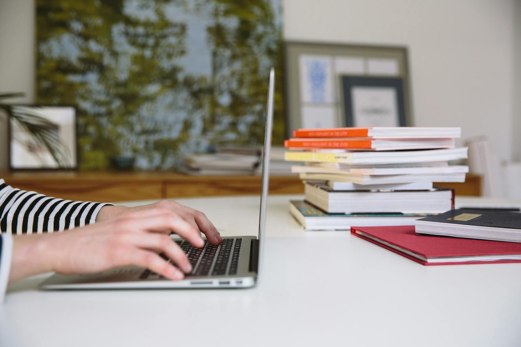 A person typing on a laptop beside stacks of books and notebooks