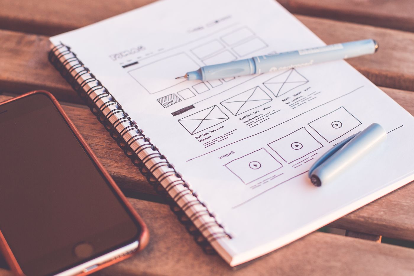 A spiral notebook with layout wireframes, a pen and a phone on a wooden table