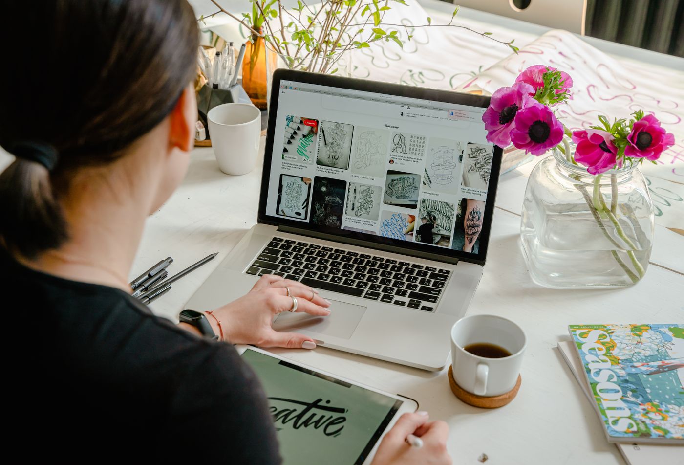 A person reviewing lettering layouts on a laptop beside flowers, a tablet and a coffee cup